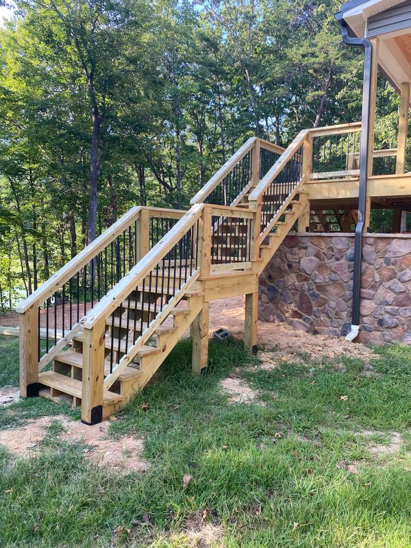 Wooden outdoor stairs with railings leading up a stone retaining wall in a grassy yard.