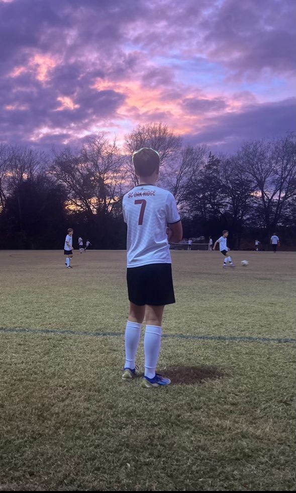 A young boy is standing on a soccer field at sunset.
