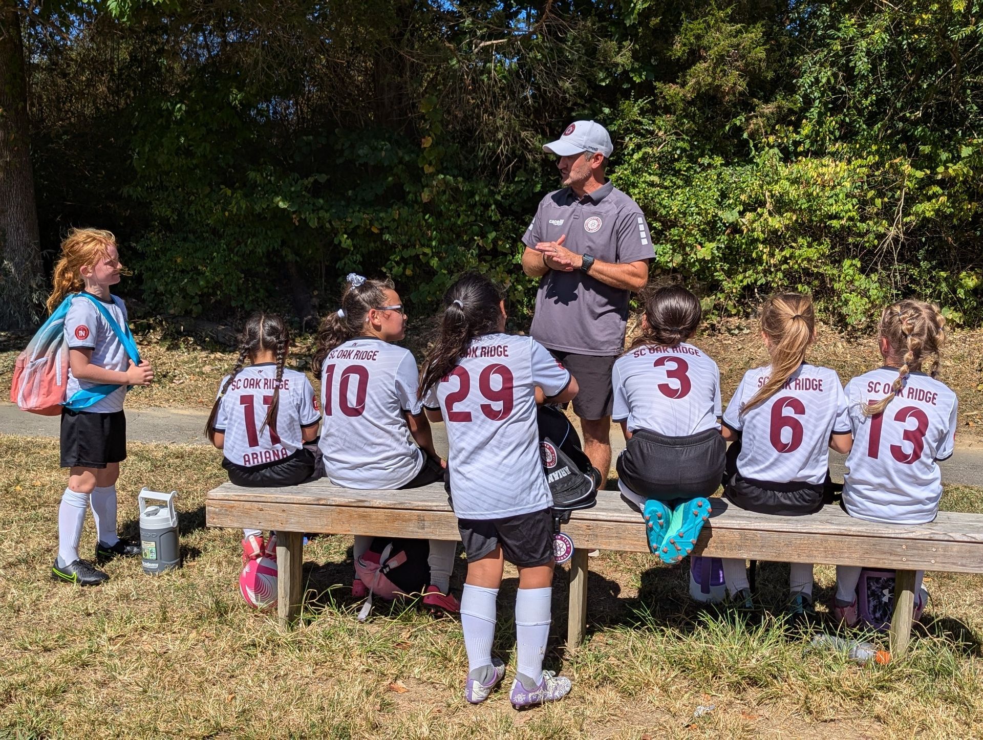 A group of young girls are sitting on a bench while a coach talks to them.