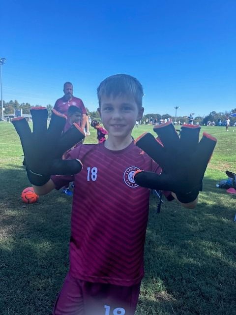 A young boy wearing a maroon shirt and black gloves is standing on a soccer field.