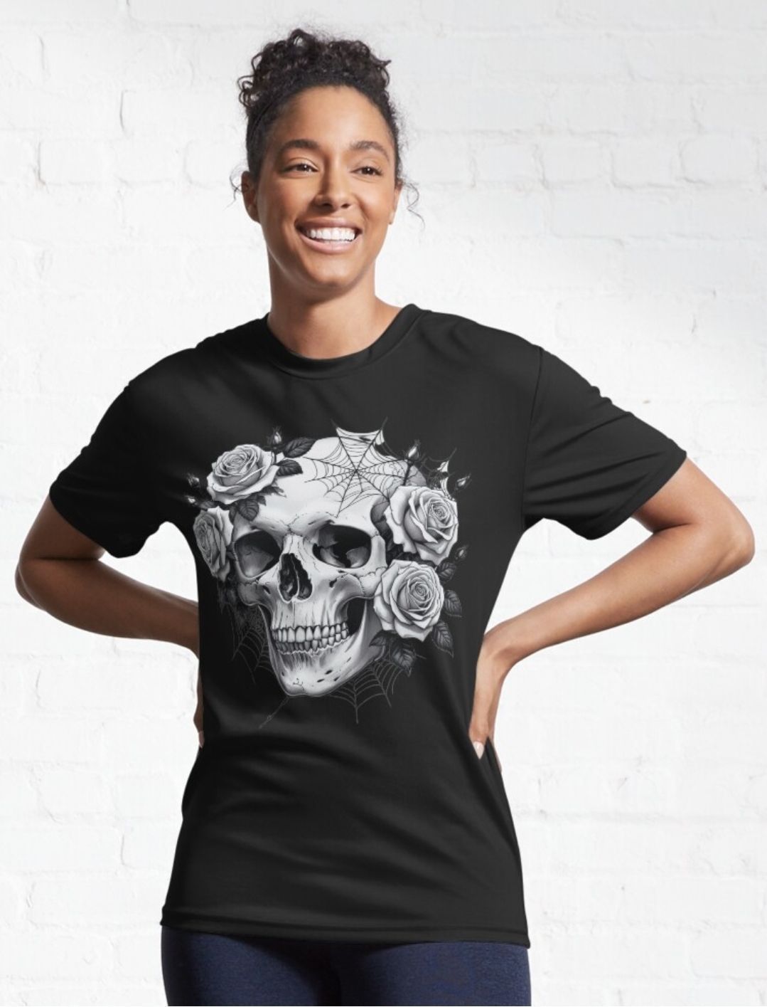 Smiling person in black T-shirt with large skull and roses graphic, standing against a white brick wall.