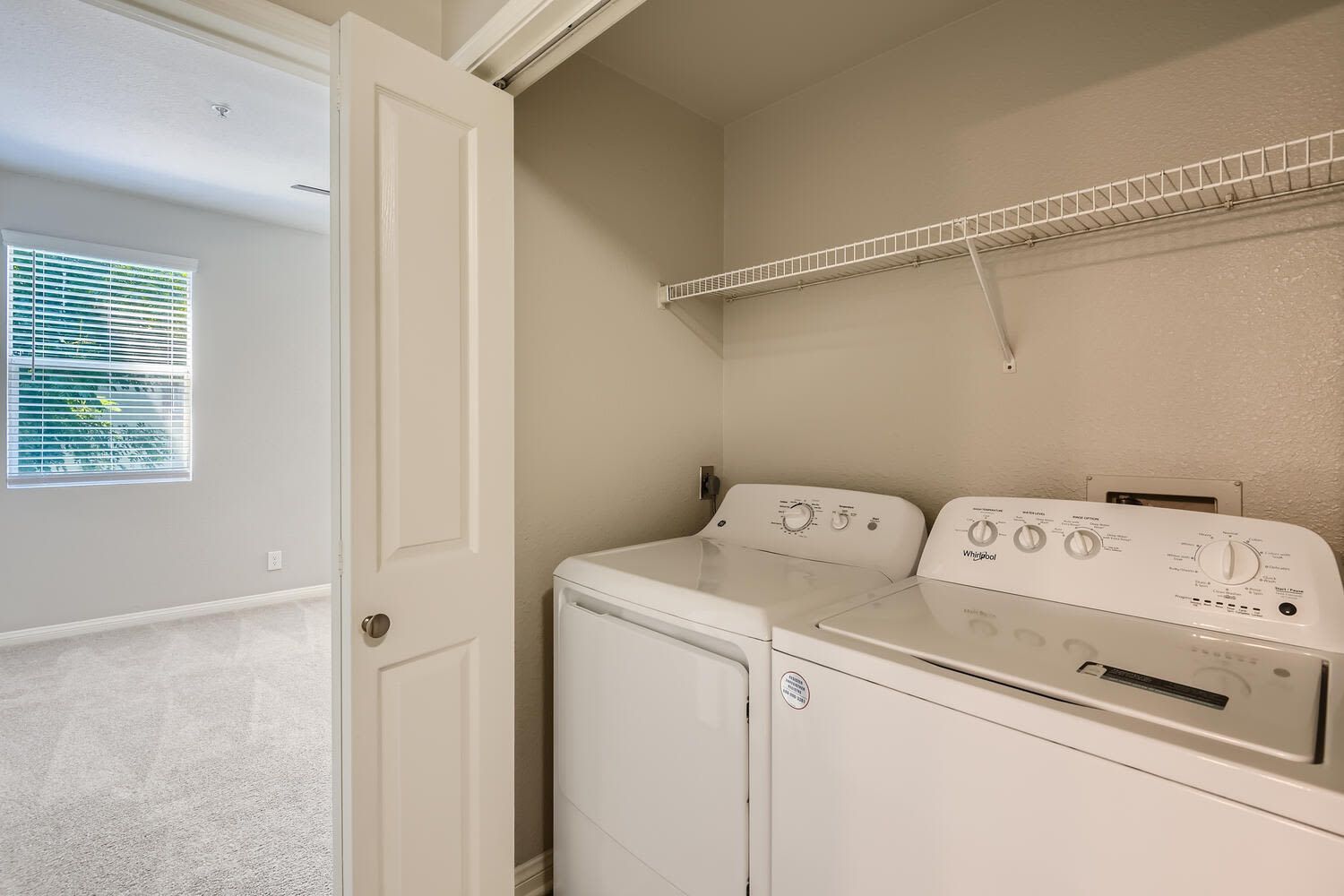Laundry closet with washer and dryer and a wire shelf above at Alicante Apartments in Aliso Viejo, CA.