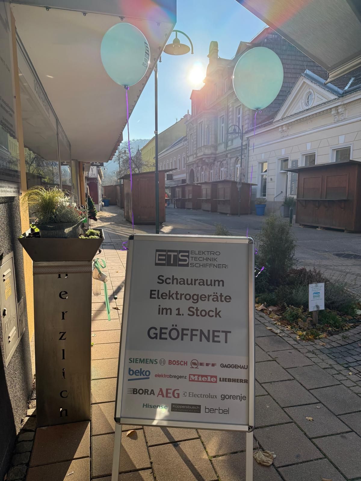 Ein A-förmiges Werbeschild wirbt für einen Ausstellungsraum für Elektrogeräte an einer sonnigen Straße, darüber hängen Luftballons.