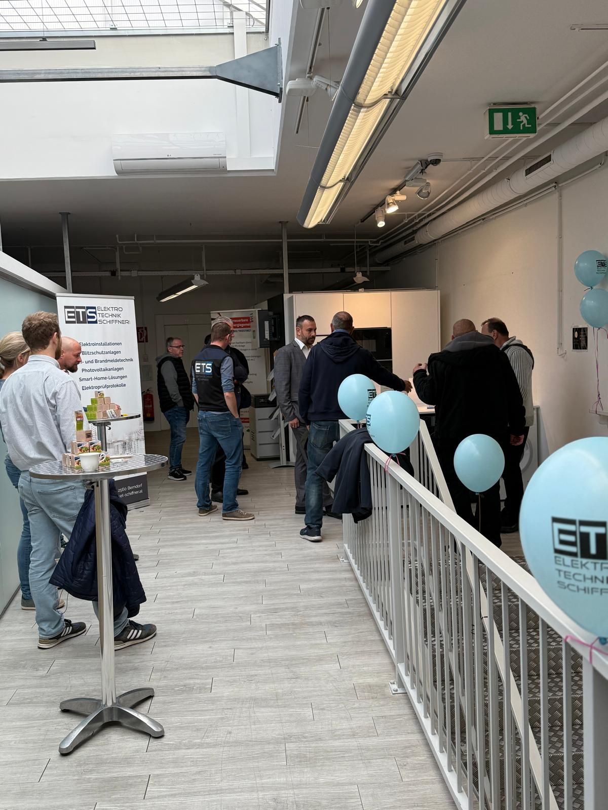 Menschen bei einer Messeveranstaltung mit einem Werbebanner, Luftballons und Ausstellungstischen in einem hell erleuchteten Flur.