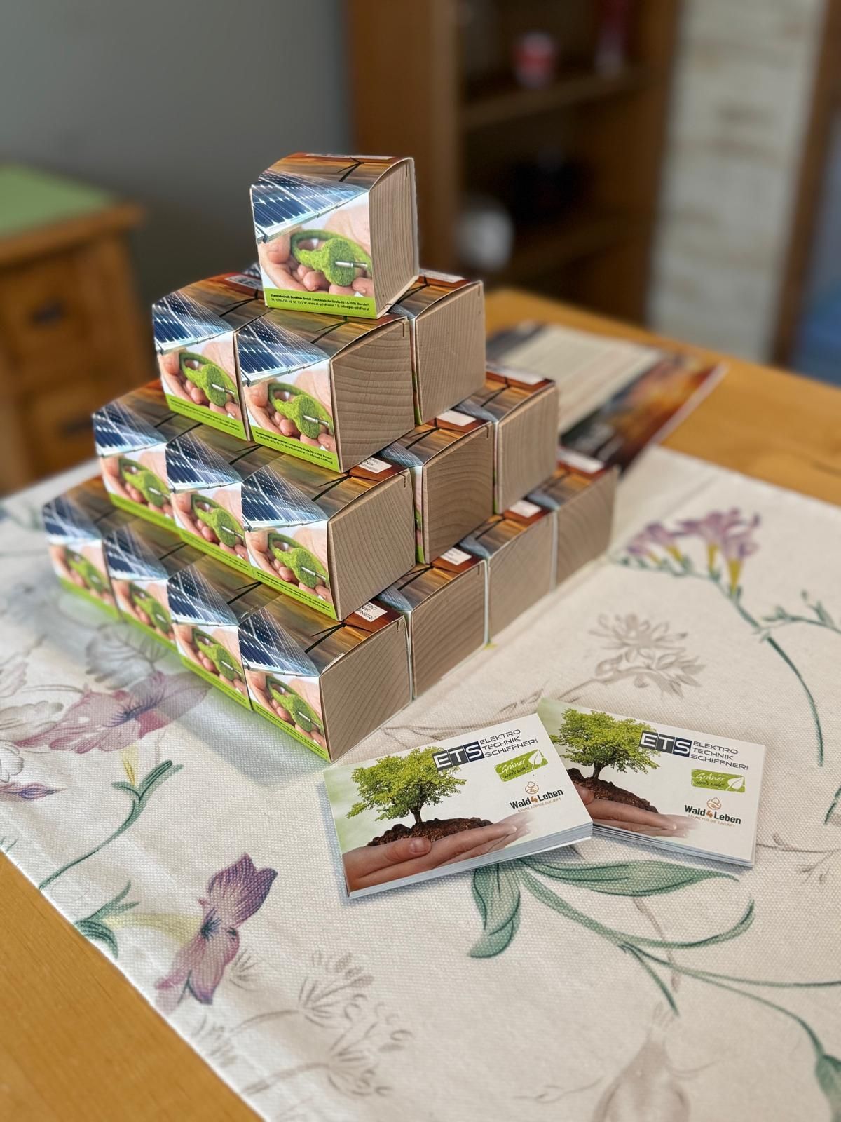 Eine Pyramide aus kleinen Pappkartons mit Pflanzenmotiven auf einer geblümten Tischdecke. Zwei ähnliche Kartons stehen neben der Pyramide.