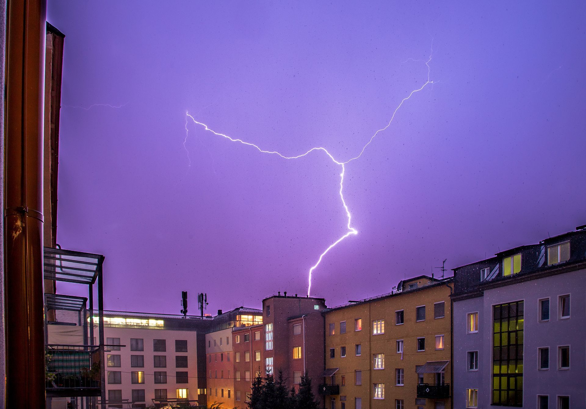 Ein heller, verzweigter Blitz schlägt über einer Reihe von Wohnhäusern vor einem tiefvioletten Dämmerungshimmel ein.