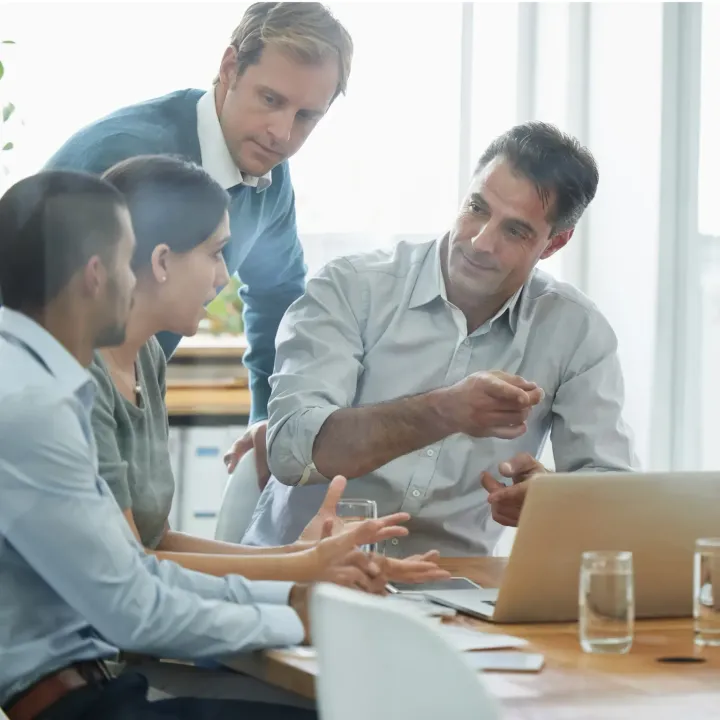 Four coworkers discuss a project around a laptop in a bright office meeting room.