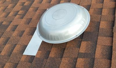 Round, silver roof vent on a brown shingle roof.