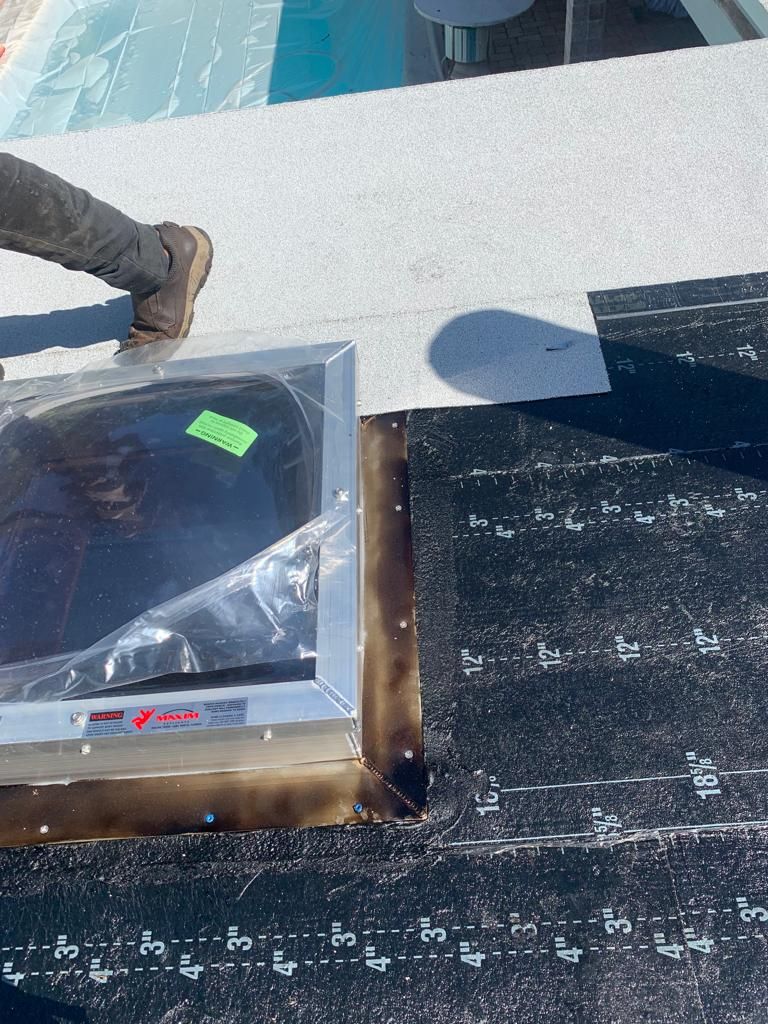 Person on rooftop installing skylight flashing. Black roofing material, silver frame.