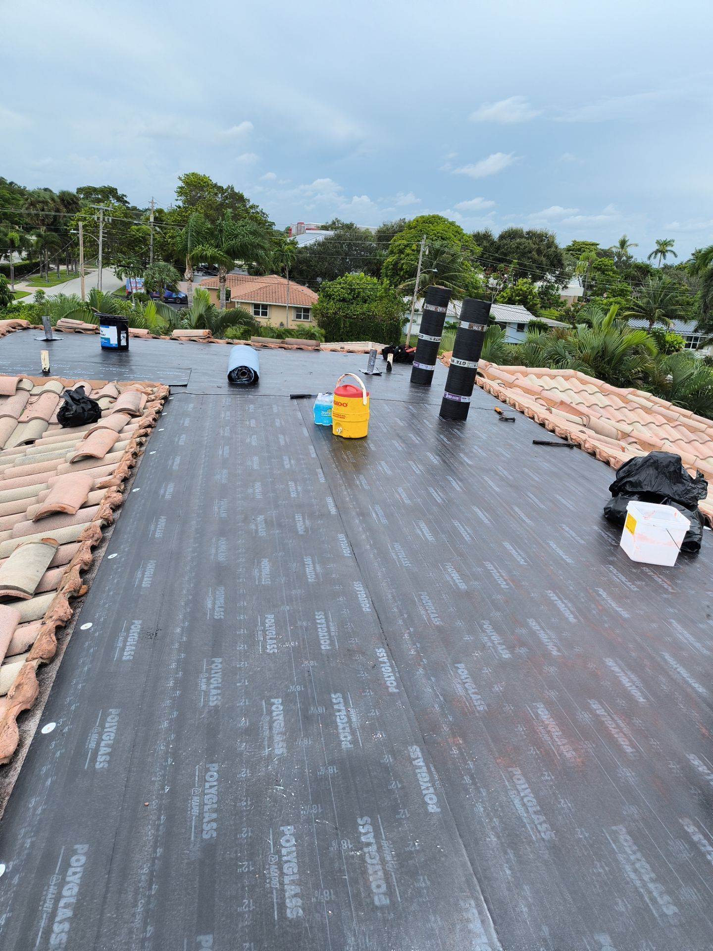 Rooftop with new dark roofing material. Various supplies, trees, and houses in the background.