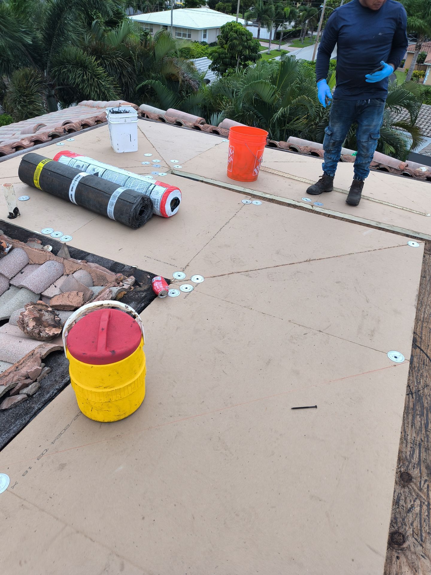 Person on a roof installing underlayment. Tan panels, buckets, and rolled materials are visible.