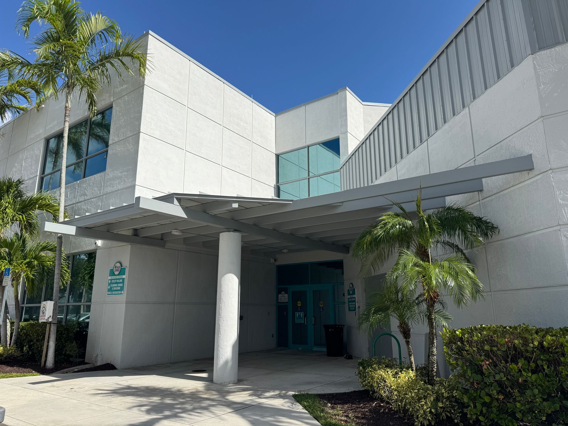 Exterior of a light-colored building with a canopy over the entrance. Palm trees and clear sky visible.