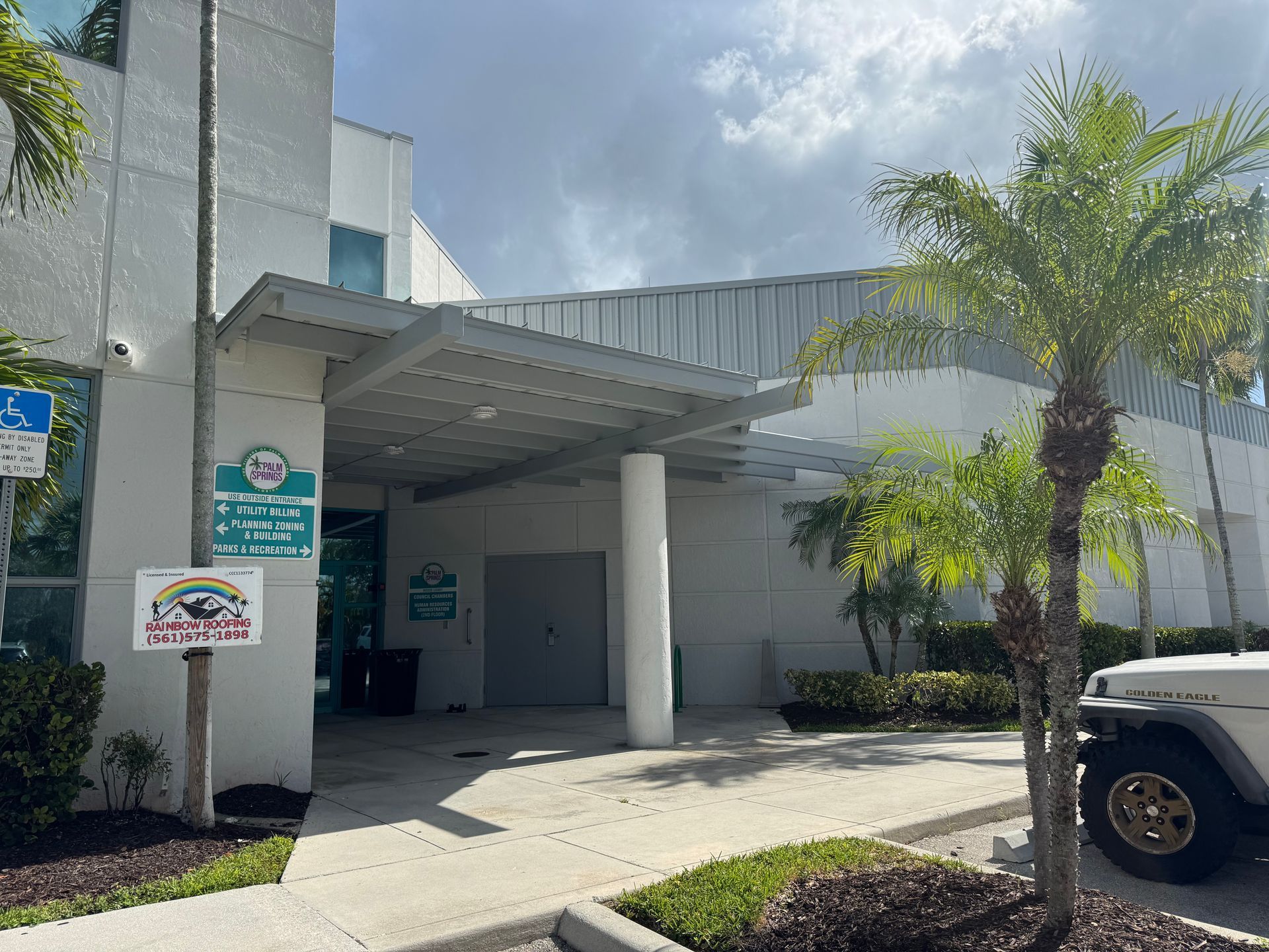 Exterior of a white building with a canopy entrance, a wheelchair access sign, and a parked Jeep.