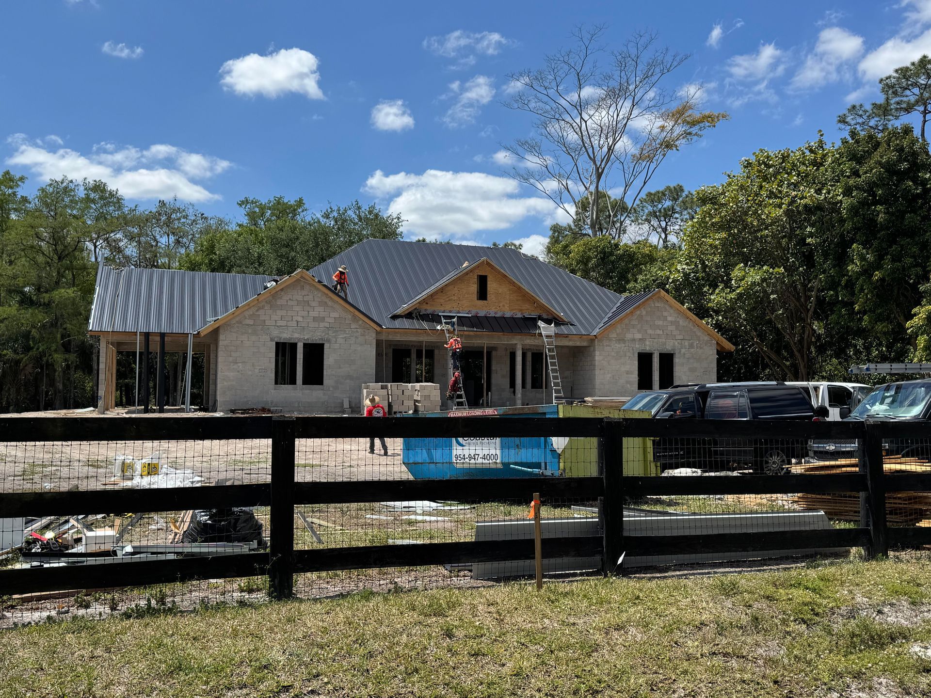 House under construction, light brick exterior, dark roof, wooden fence, blue sky, surrounded by trees.