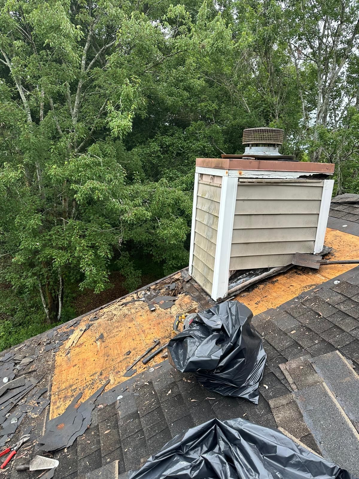 Damaged roof with a chimney. Black trash bags sit on the shingles. Lush green trees in the background.