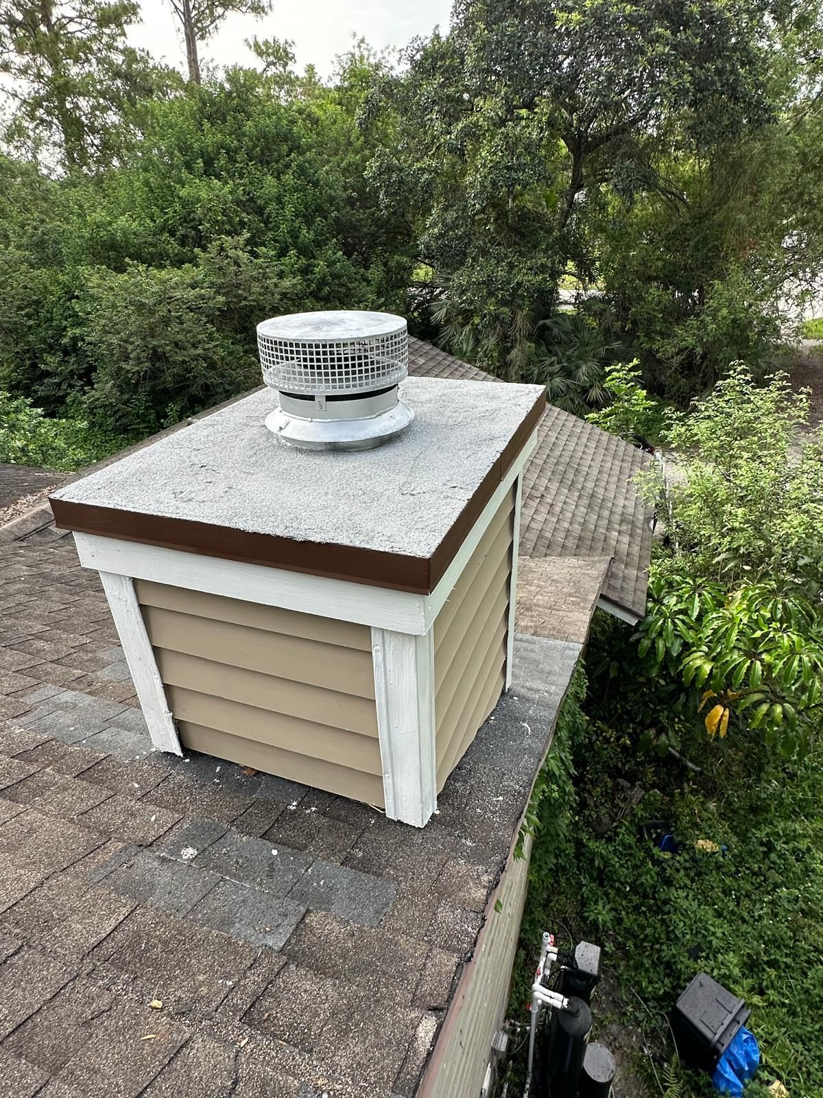 A roof vent on a tan-sided box, brown trim, and a shingle roof. Surrounded by trees.