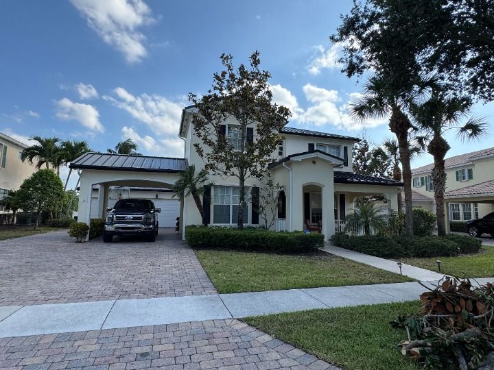 Two-story cream-colored house with a black car in the driveway, surrounded by palm trees and green grass.