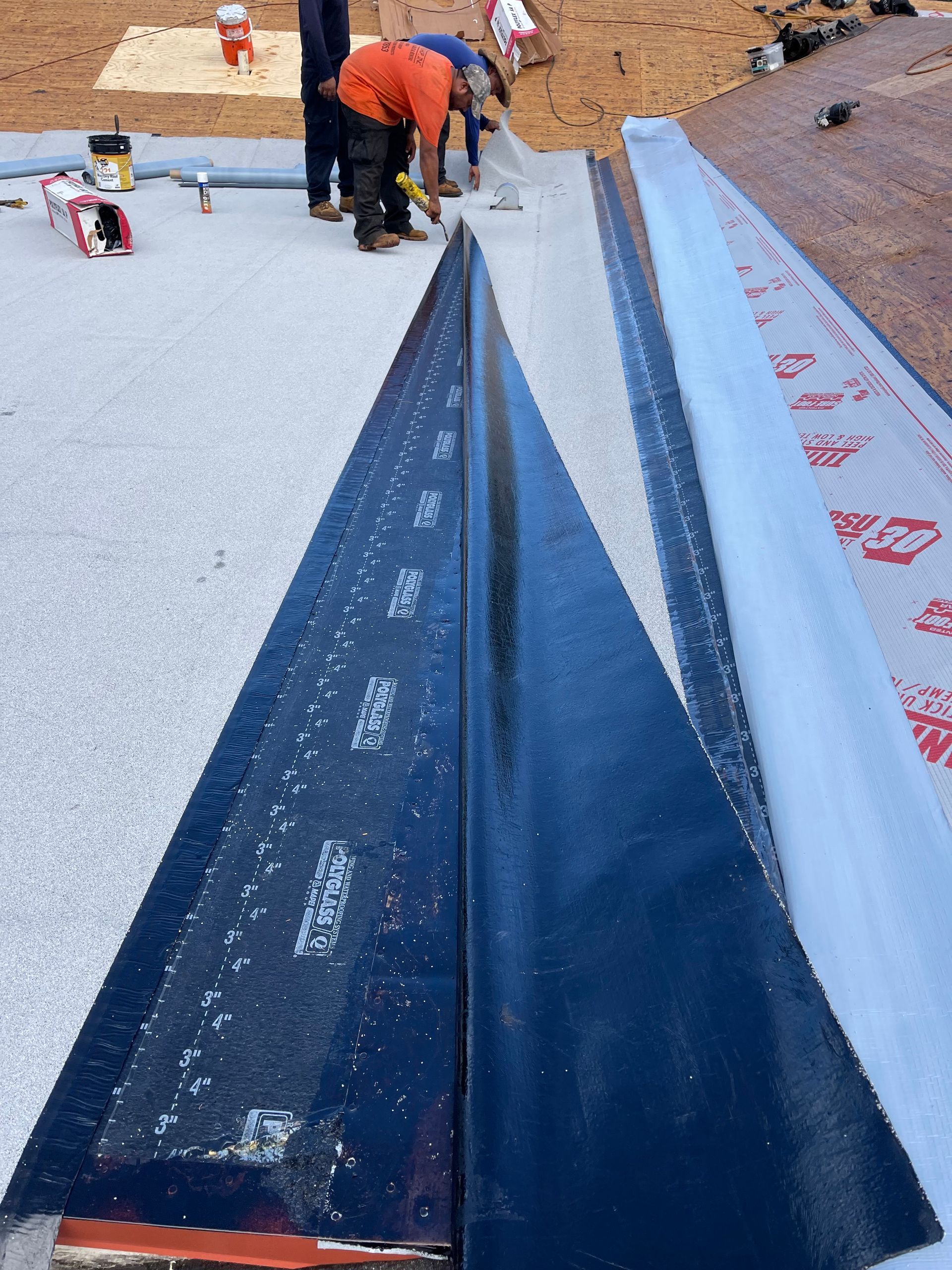 Workers installing roofing material, dark strip in a trough, blue/gray surface, sunny outdoor setting.