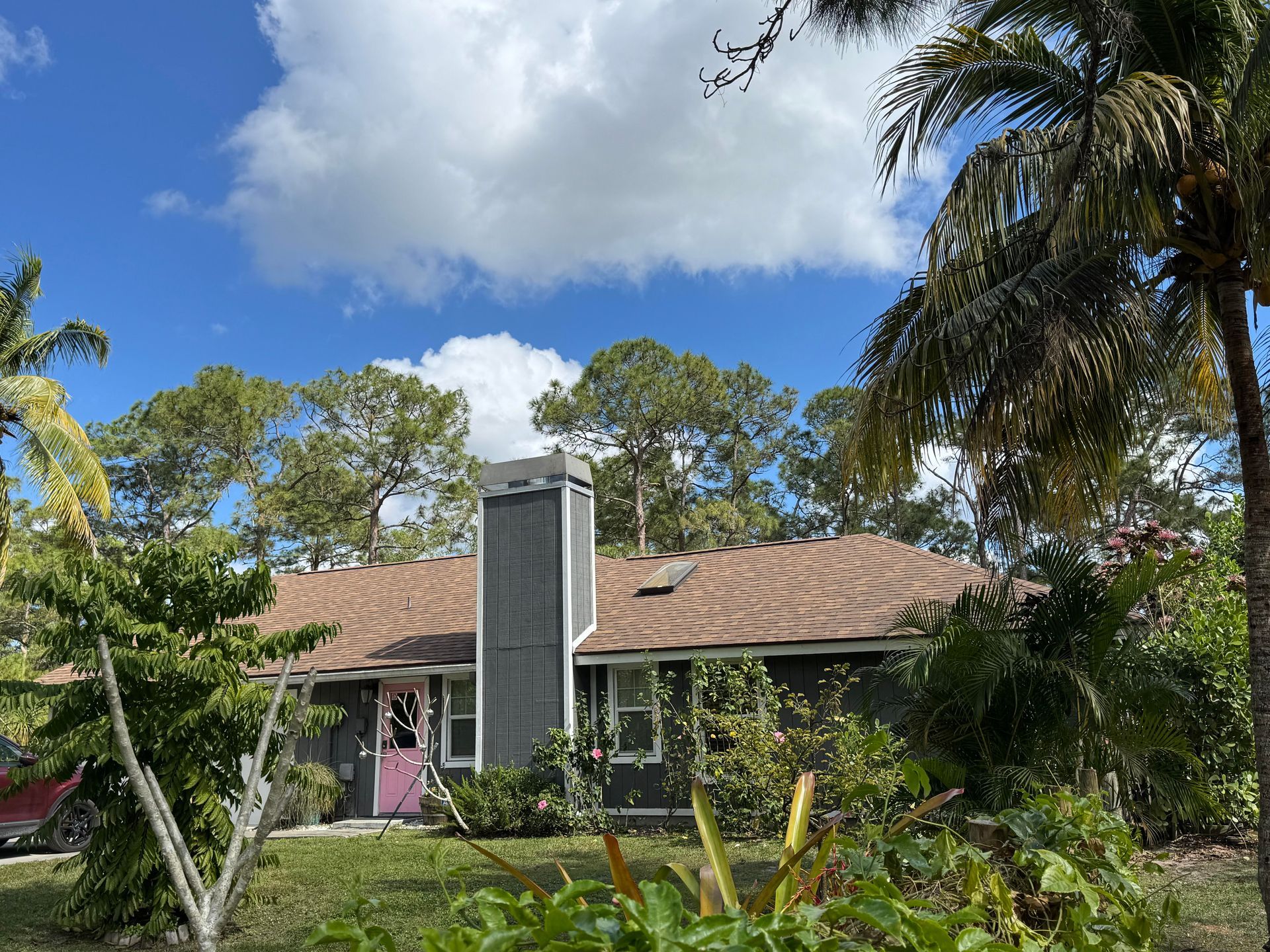 A gray house with a brown roof and a tall gray chimney, surrounded by trees and greenery.