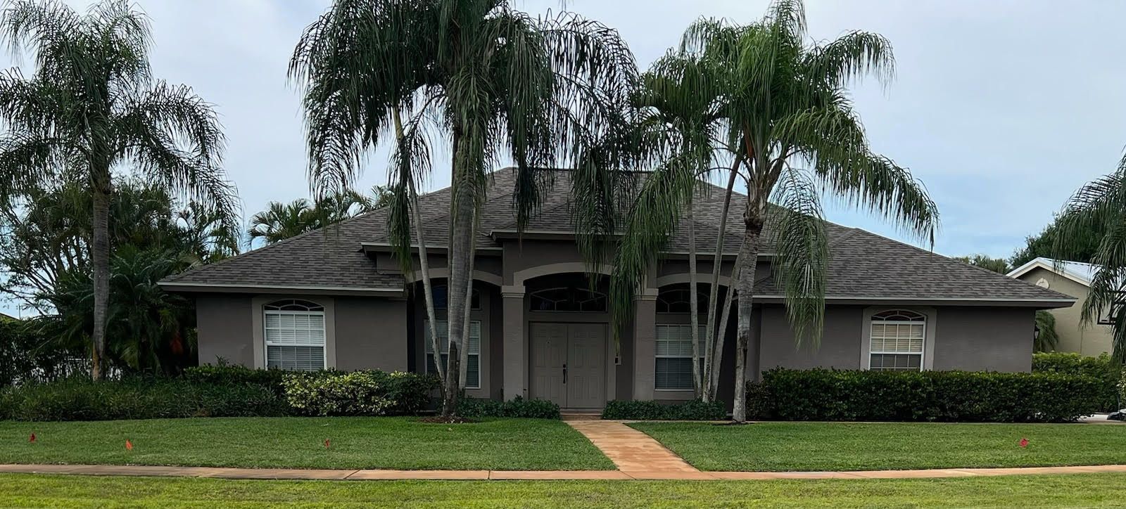 A beige house with a grey roof, palm trees, and a brick pathway under an overcast sky.