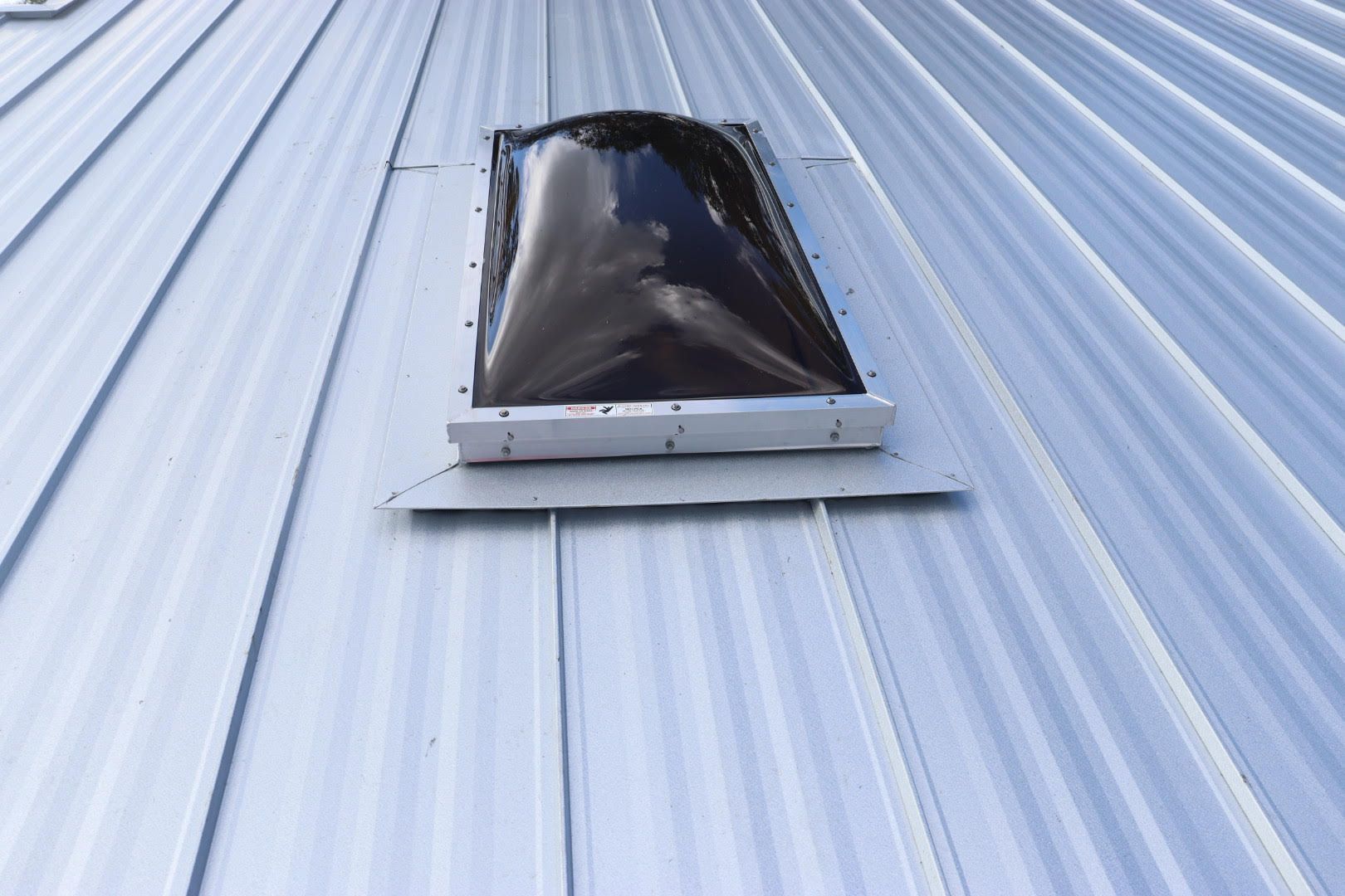 Skylight on a corrugated metal roof. Black plastic bubble with a silver metal flashing.