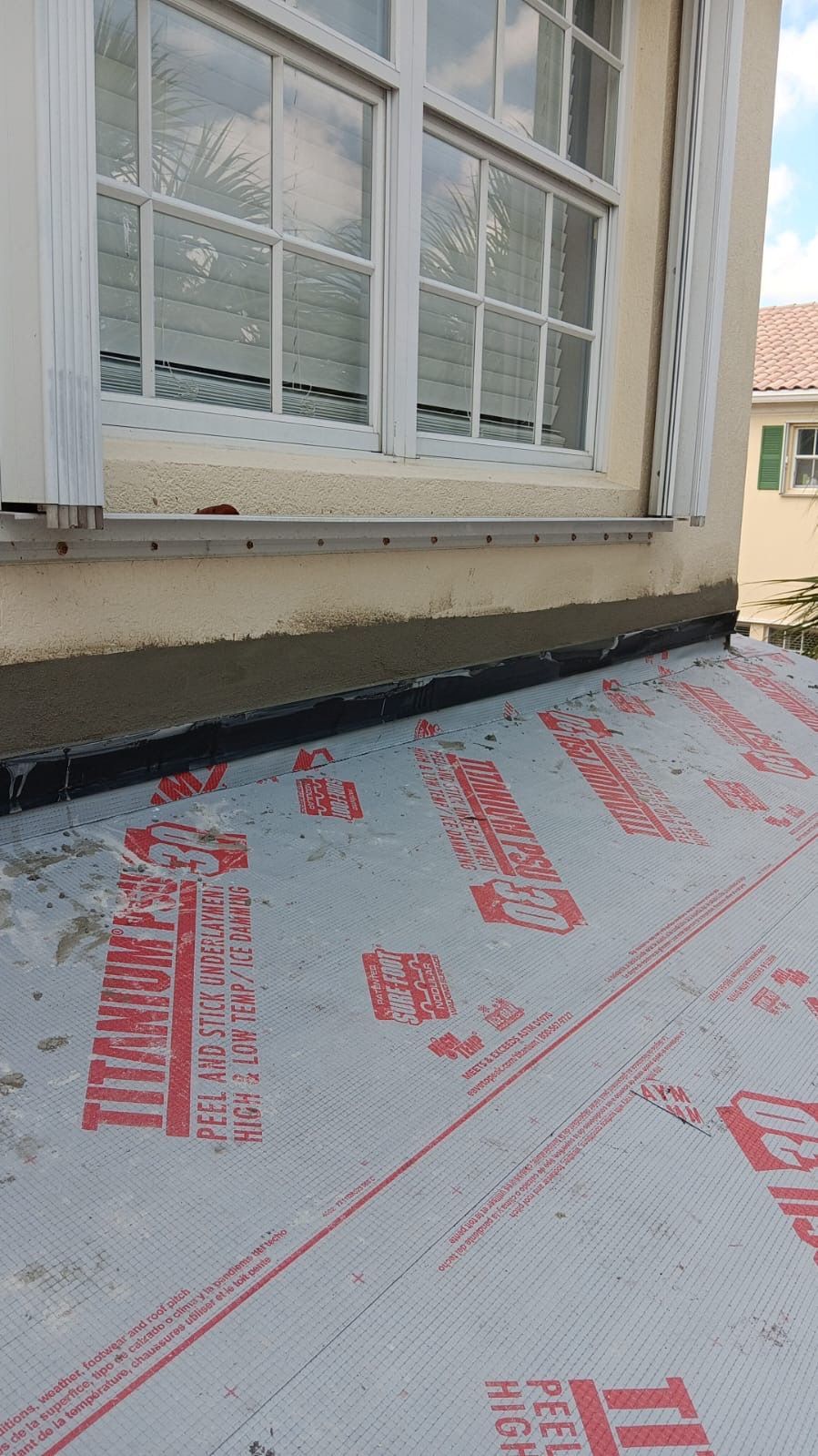 Close-up of a roof with underlayment and a window above. Beige building, window, and white underlayment.