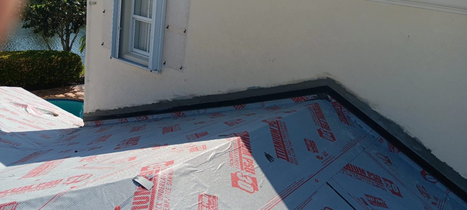 Roofing materials being installed at the corner of a building; white walls, blue protective covering, and window.