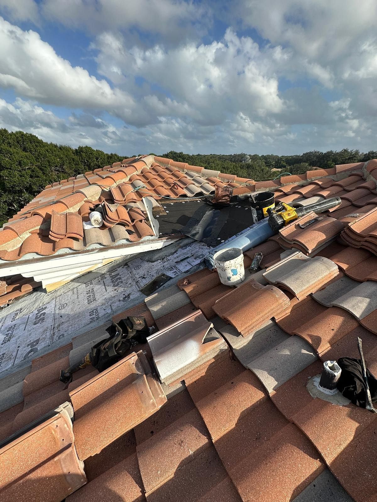 Rooftop with missing tiles exposing underlayment. Blue sky and trees in the background. Construction underway.