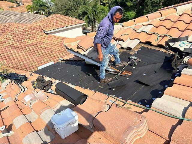 Roofer repairs a tile roof, standing on black underlayment. Bright sun.