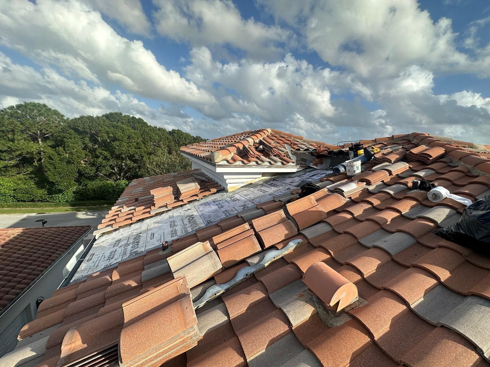 Rooftop with broken terracotta tiles being repaired, blue sky, and trees in the background.