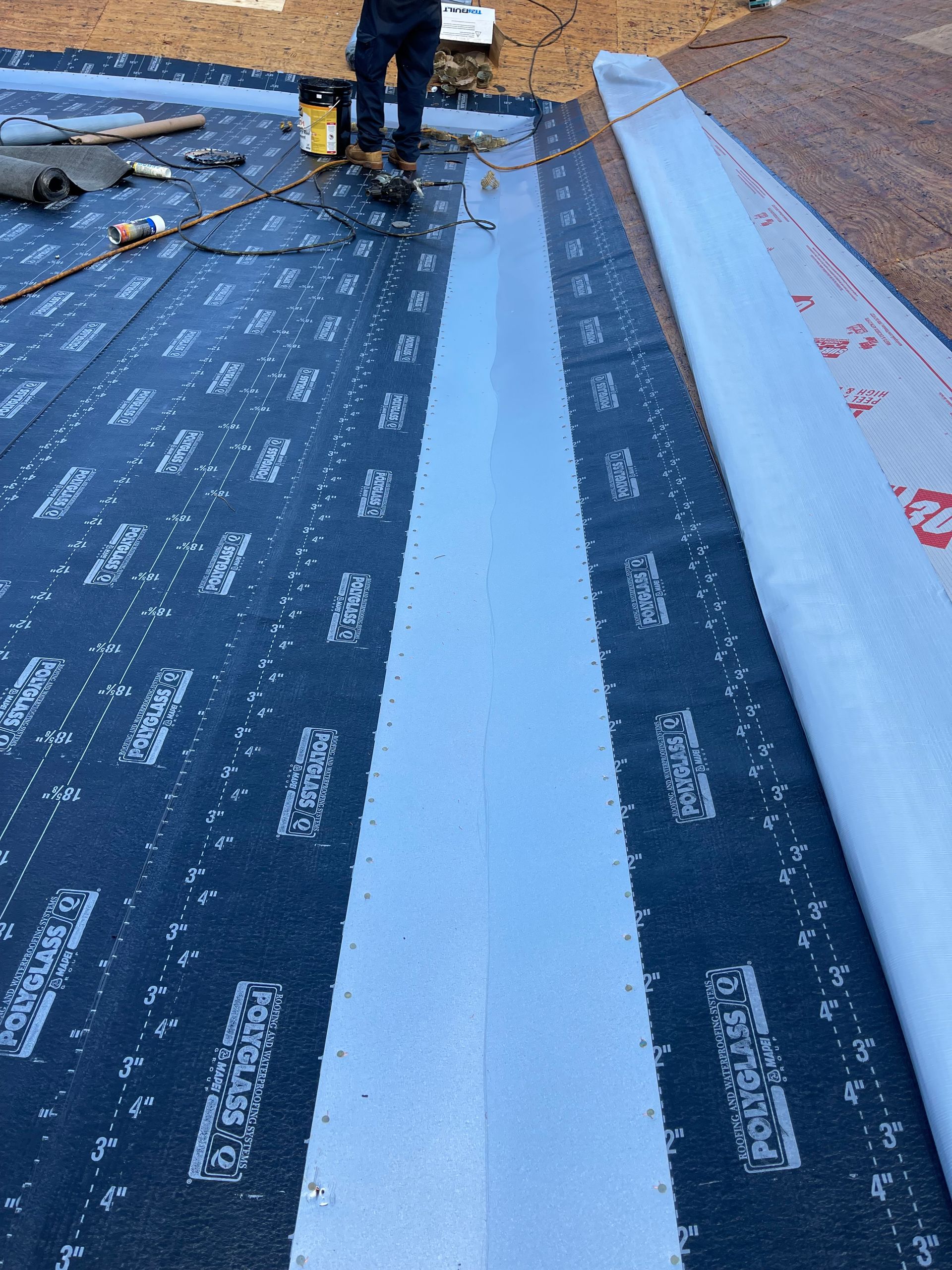 Person roofing; applying sealant to seam of rolled roofing material. Black material, blue sealant.