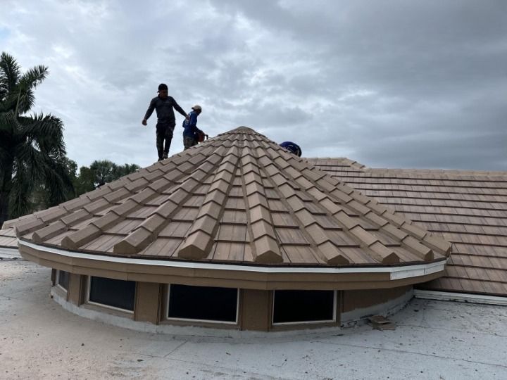 Workers installing brown tiles on a rounded roof with skylights against a cloudy sky.