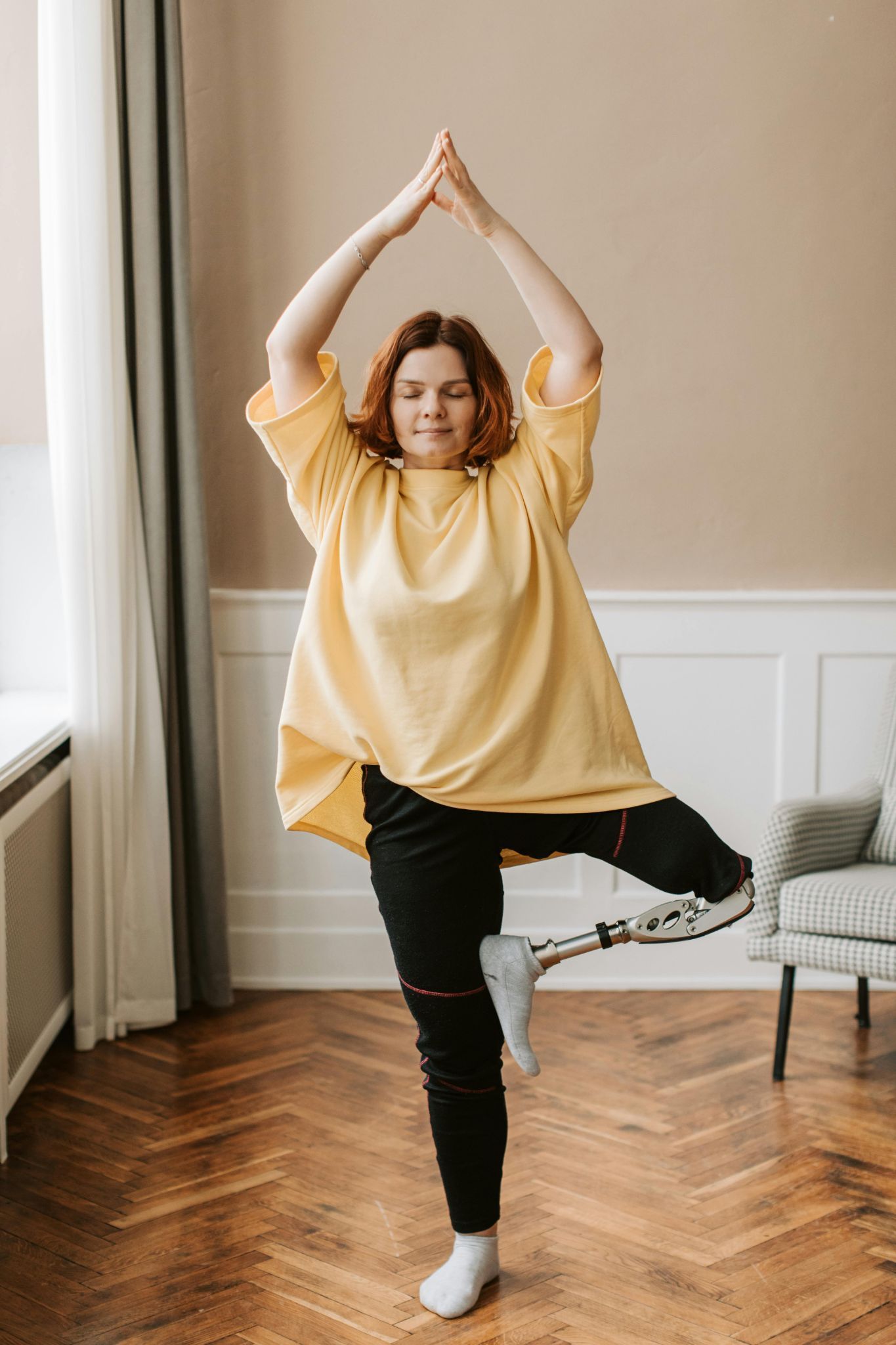 Woman balancing in yoga tree pose indoors near a window, wearing a yellow top and black pants.