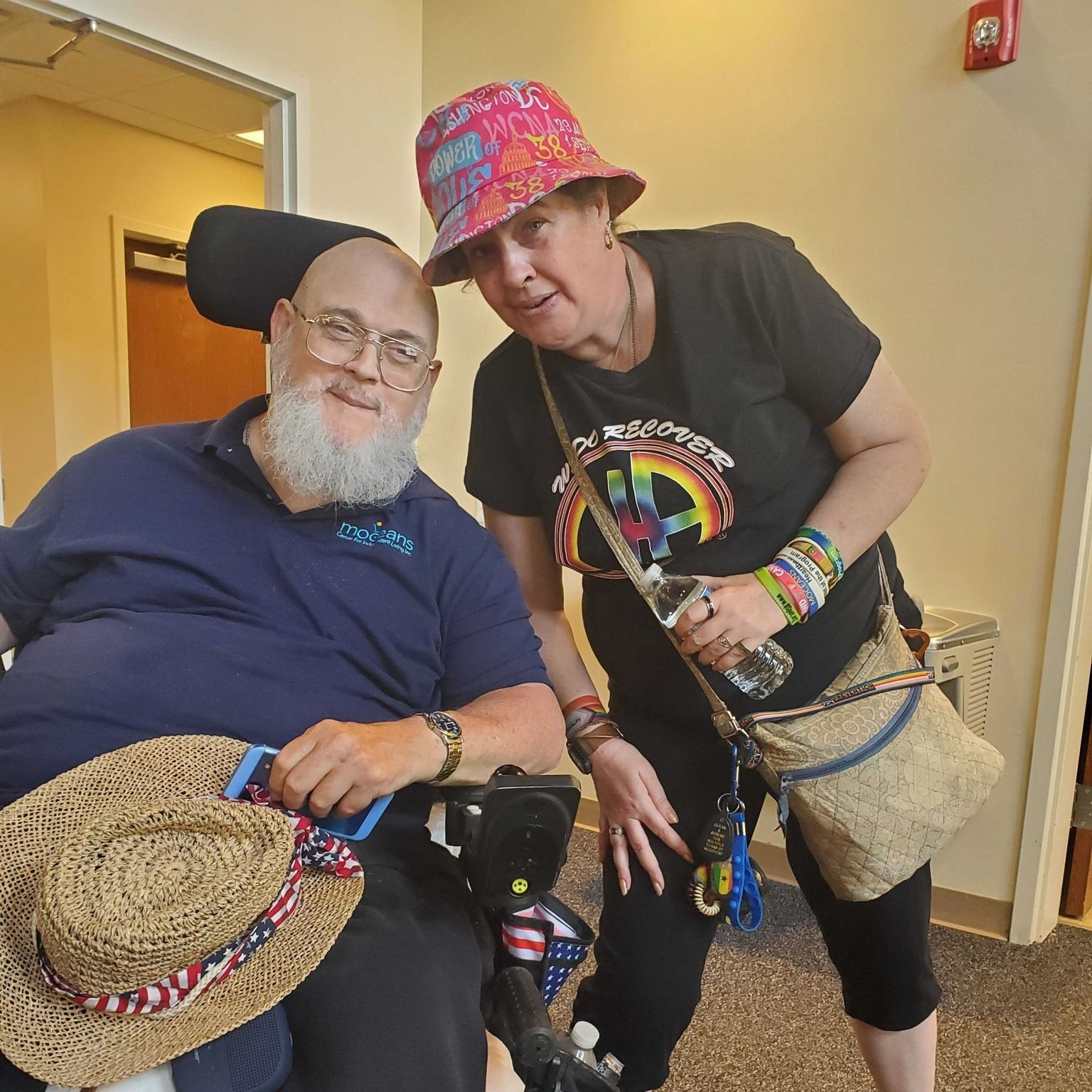 Two people smiling indoors, wearing colorful hats and holding woven hats and a cane.