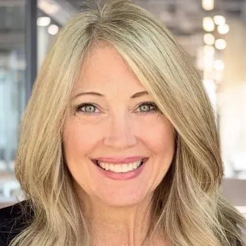 Woman with blonde hair smiling at the camera, wearing a black shirt, in a brightly lit setting.