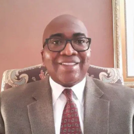 Man in suit and glasses smiling, seated in a decorated chair, against a peach-colored wall.