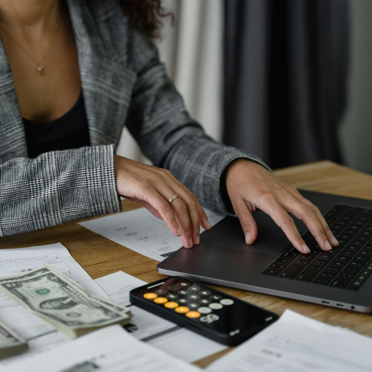 Person in blazer using a laptop, calculator, and cash on a table covered in papers.