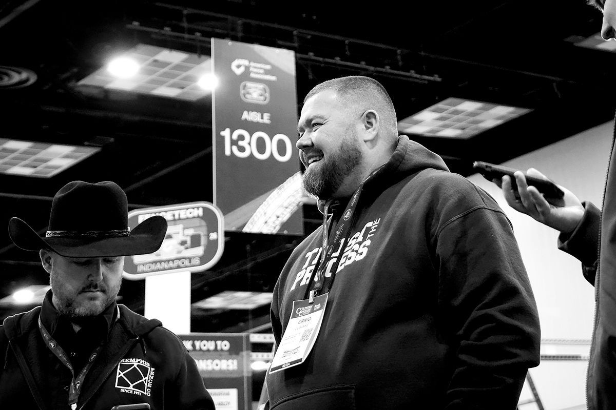 Two men talking at a trade show booth, with signage and one man smiling in a dark hoodie.