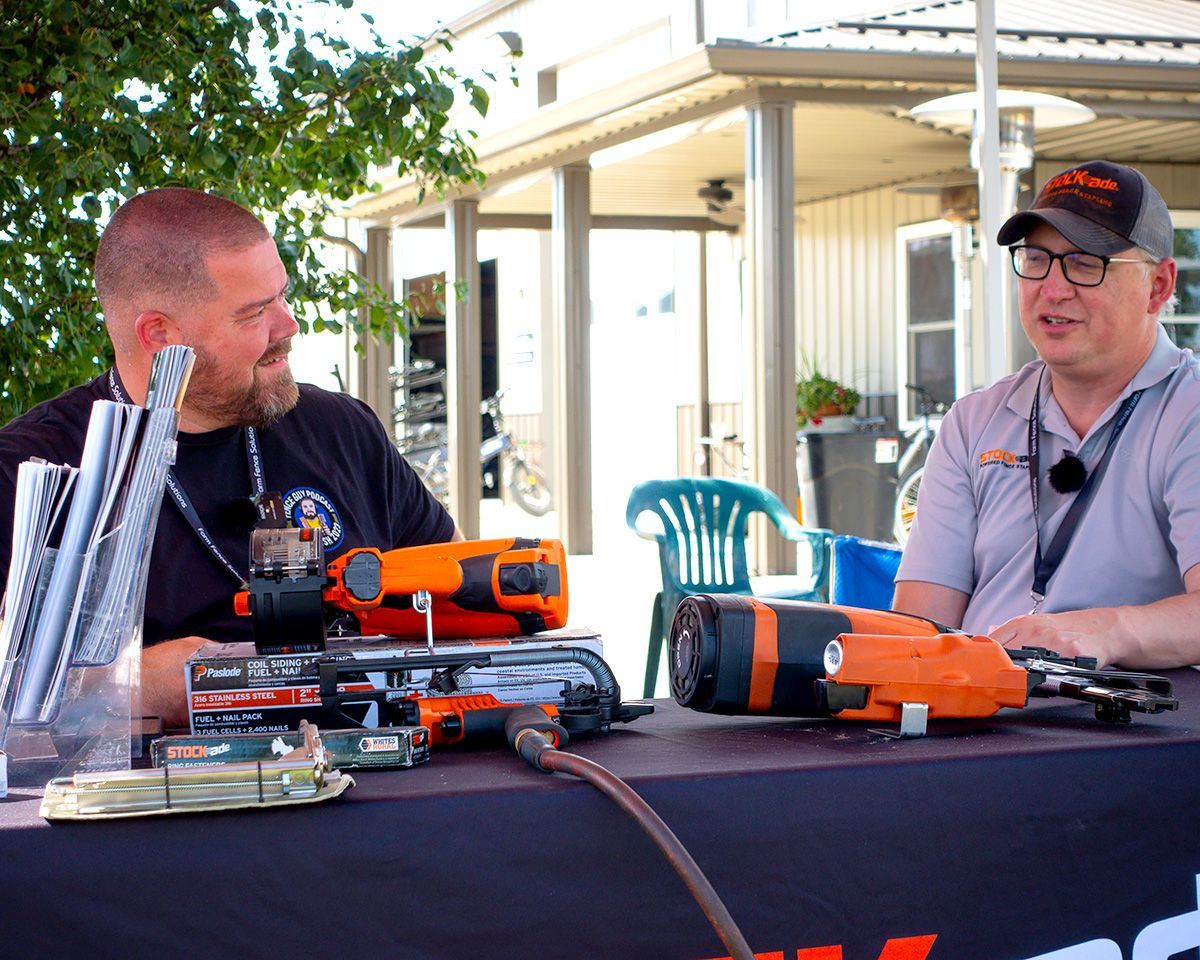 Two men at a table displaying orange power tools outdoors under a canopy