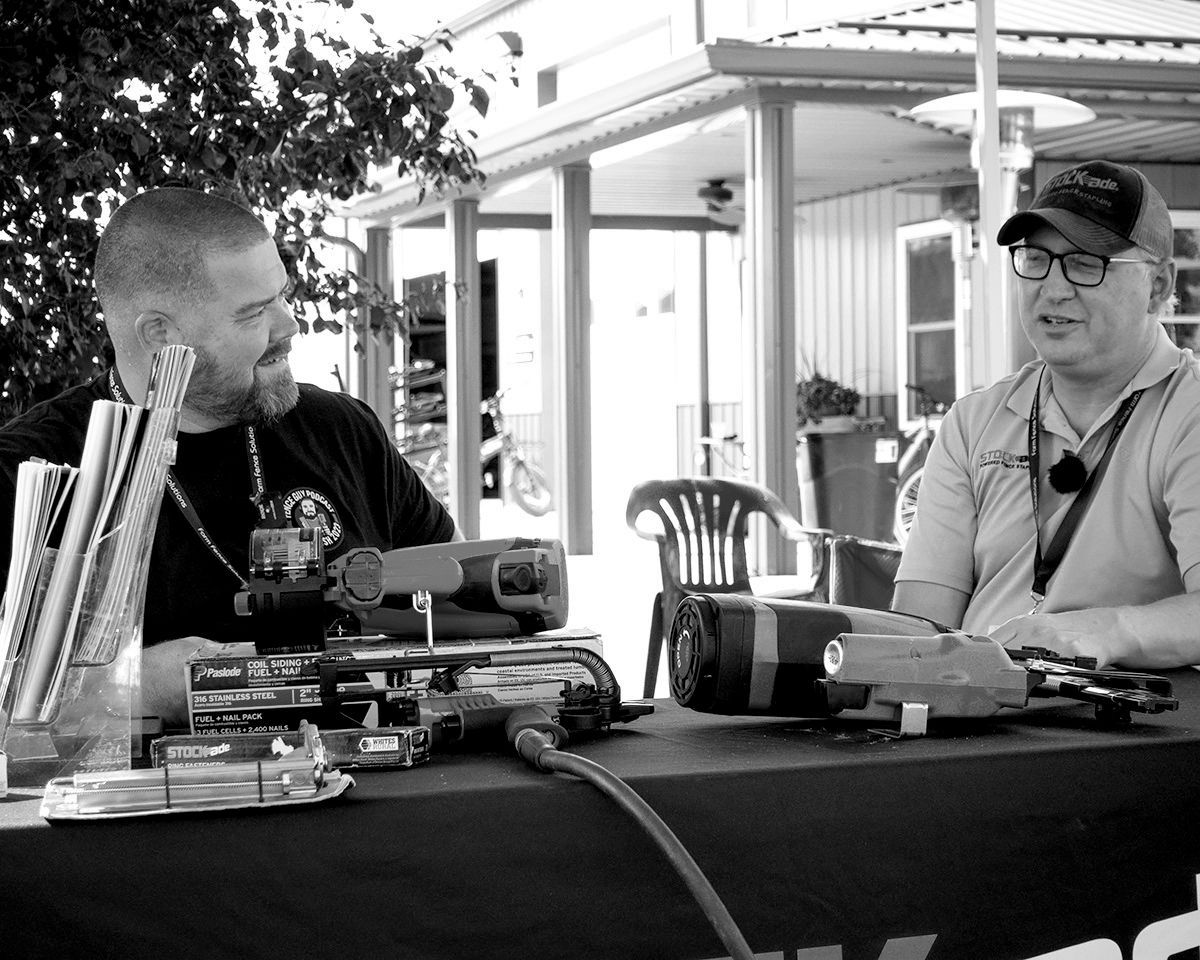 Two people sit at a table with cameras and equipment, talking outdoors under a shaded patio.