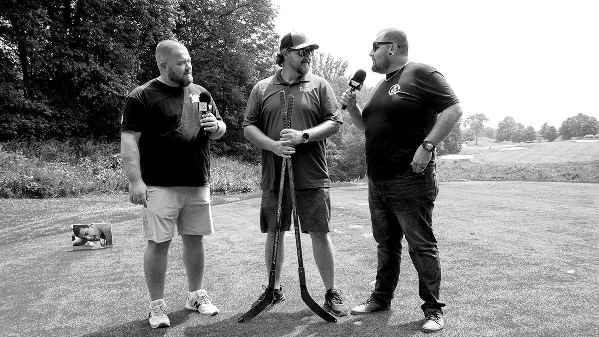 Three people standing on a grassy field, holding microphones and speaking outdoors.