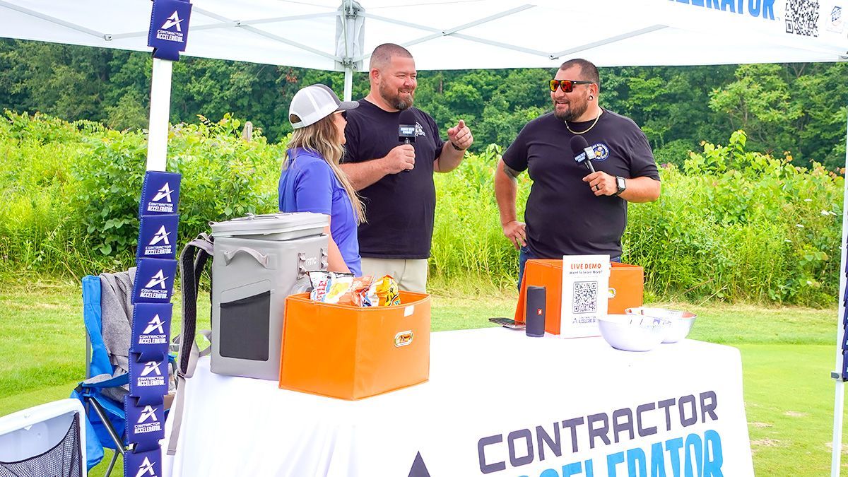 Two men speaking at an outdoor contractor expo booth beside branded displays and an orange cooler