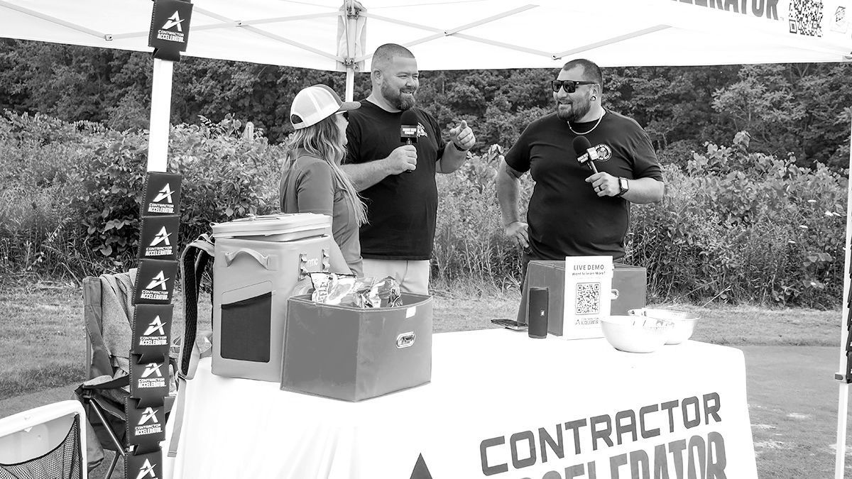 Two men at an outdoor contractor expo table with promotional items and a banner.