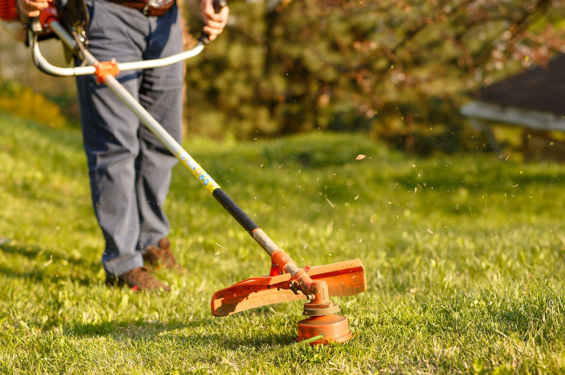 Worker in gray pants trimming green grass with an orange string trimmer in sunlight.