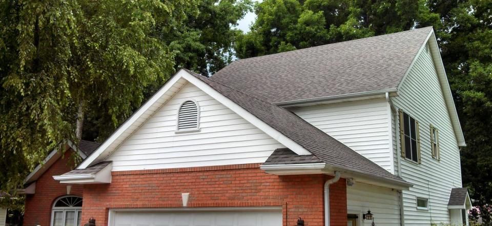 White and red two-story house with a garage, gray shingled roof, and trees in the background