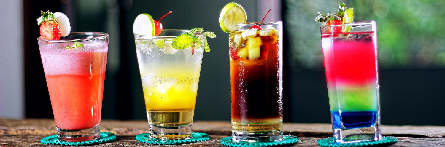 Four colorful cocktails in tall glasses lined up on a wooden table with fruit garnishes