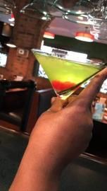 Hand holding a martini glass with a green and red layered drink in a bar setting