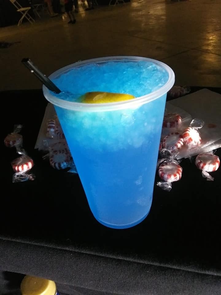 Blue frozen drink in a clear cup with a lemon wedge and straw on a dark table with candy lights.