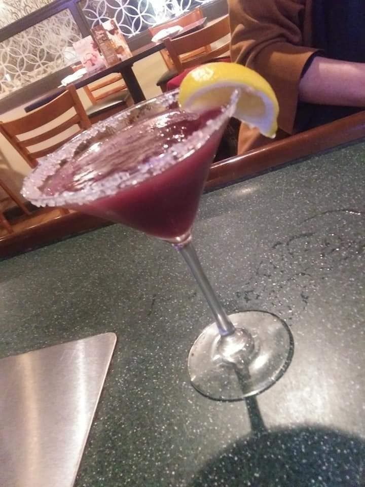 A purple cocktail in a martini glass with a lemon slice on a restaurant table.