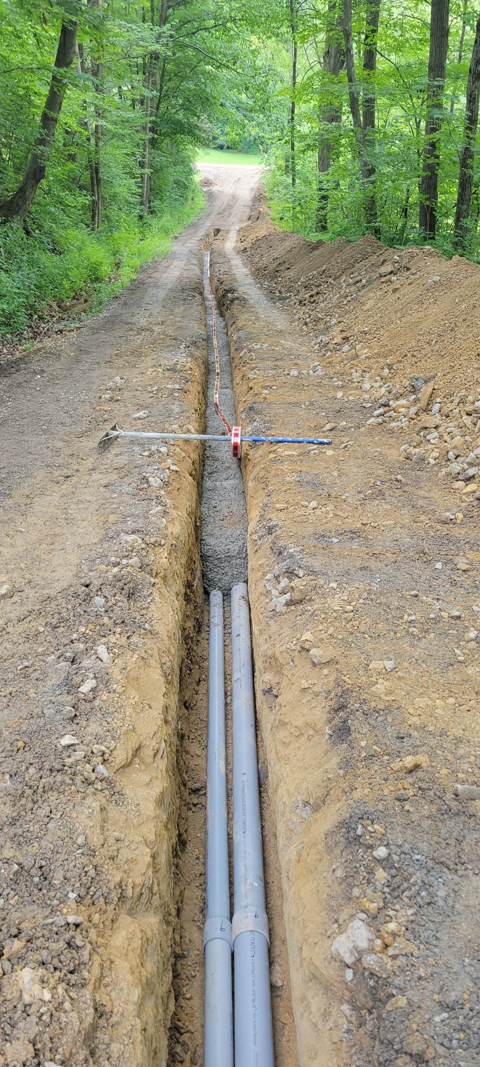 Trench in a dirt path with grey pipes. The path is in a forest with green trees on both sides.